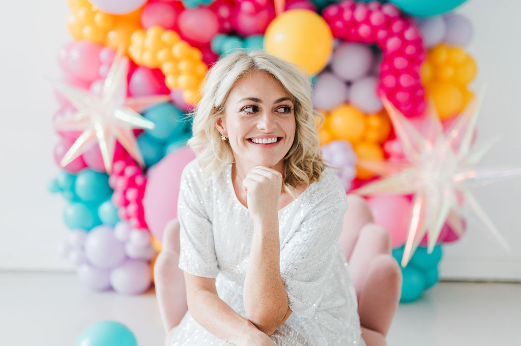 Brand photo of a North Texas balloon artist sitting in a pink chair in front of a balloon wall she designed.