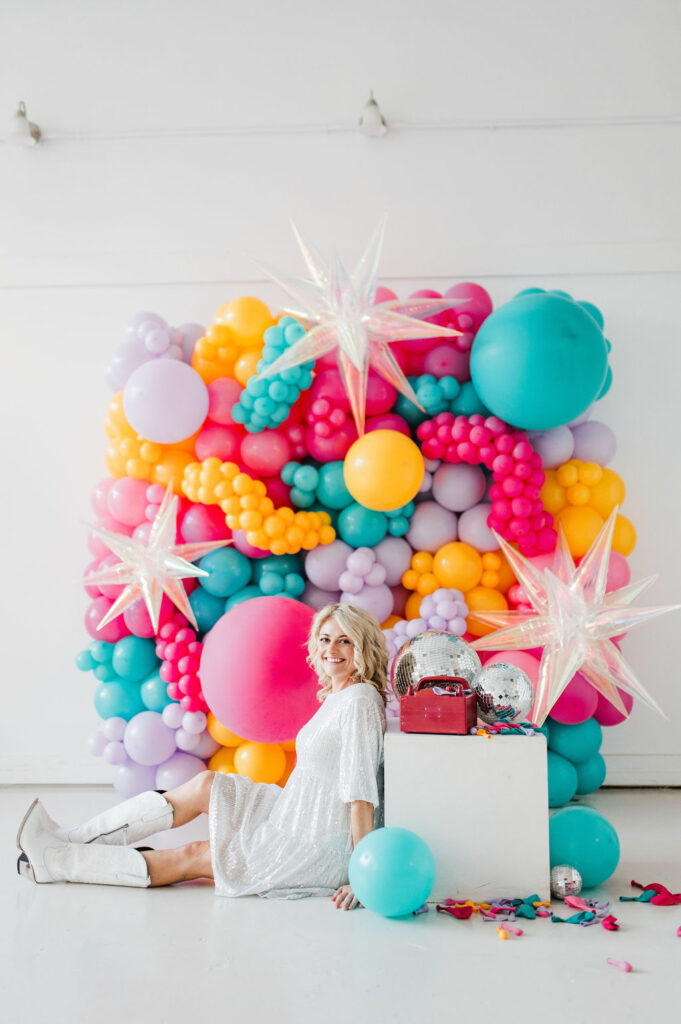 Brand photo of a North Texas balloon artist sitting in front of a balloon wall she designed.