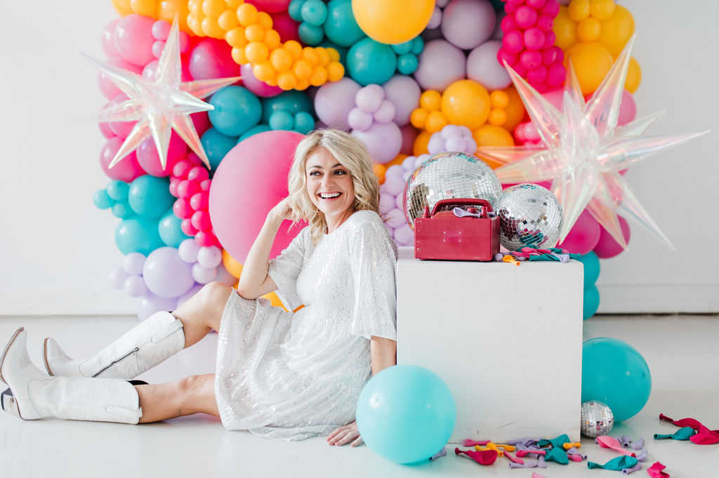 Brand photo of a North Texas balloon artist sitting in front of a balloon wall she designed.