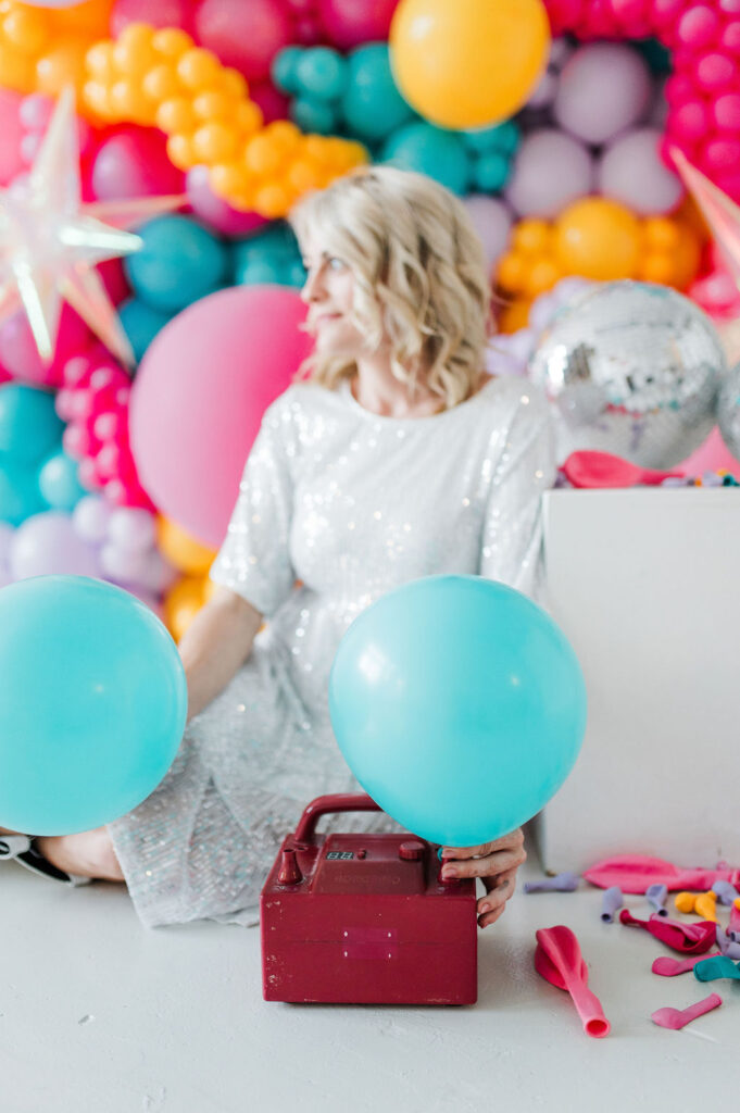 Brand photo of a North Texas balloon artist sitting in front of a balloon wall she designed.