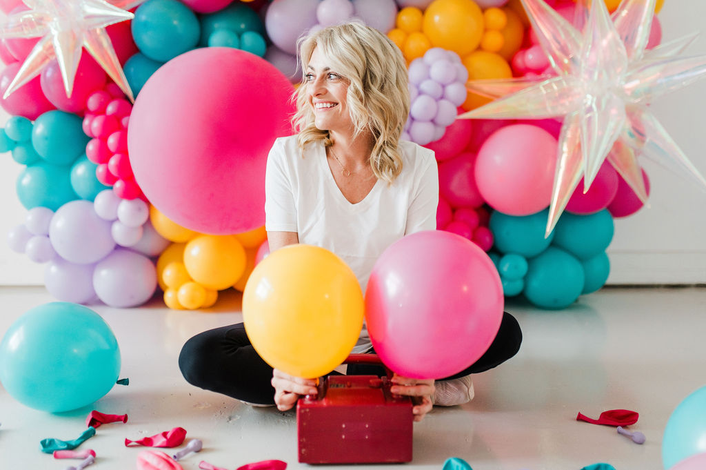 Brand photo of a North Texas balloon artist sitting in front of a balloon wall she designed.