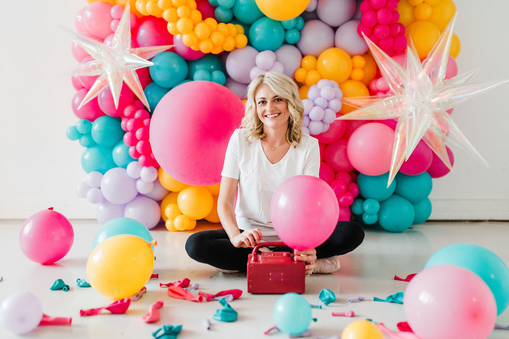 Brand photo of a North Texas balloon artist sitting in front of a balloon wall she designed.