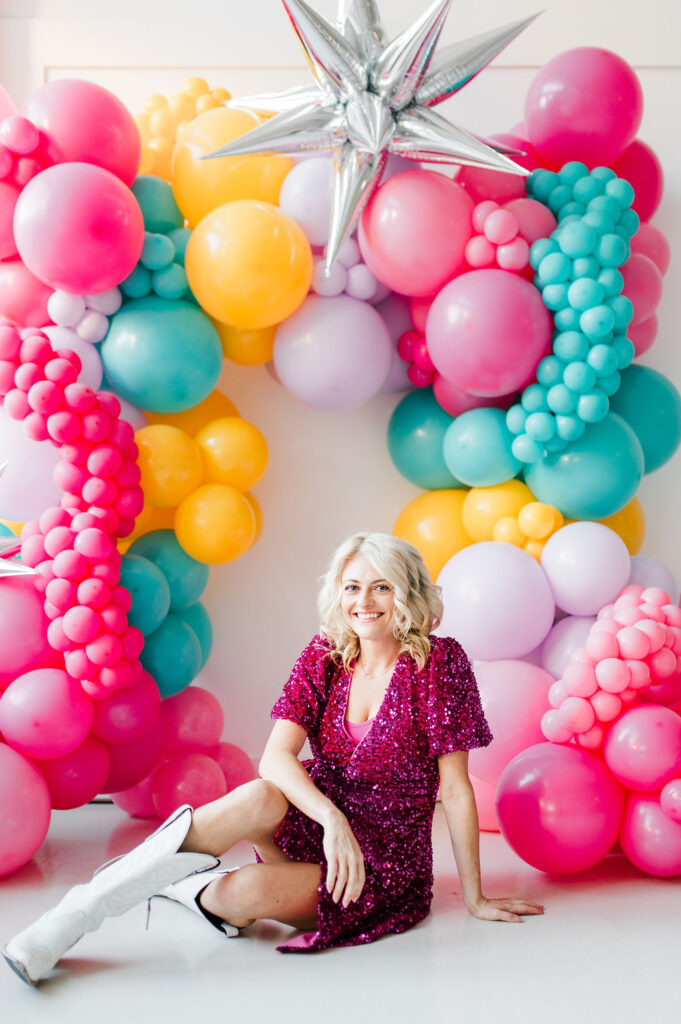 Brand photo of a North Texas balloon artist sitting in front of a balloon wall she designed.