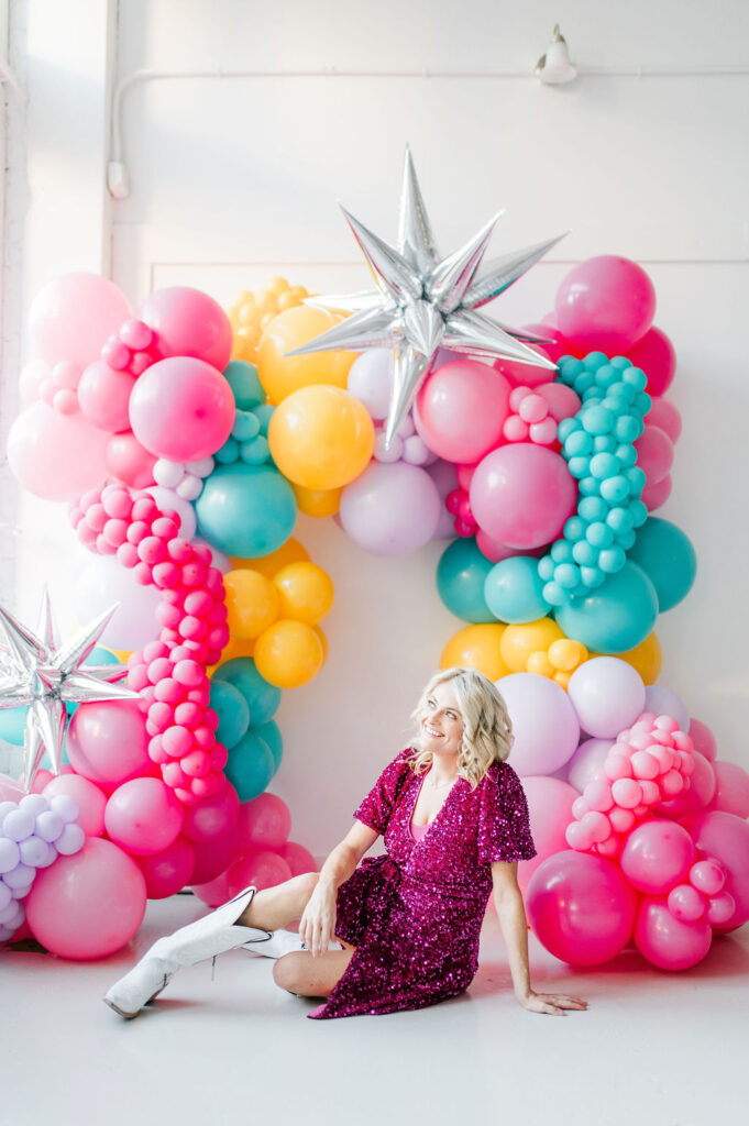 Brand photo of a North Texas balloon artist sitting in front of a balloon wall she designed.